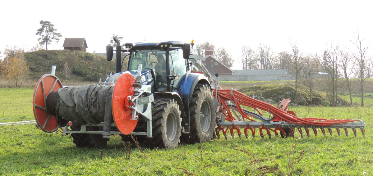 Bild på traktor med slangvinda fram och spridare med spridningsramp bak på ett fält med vall.