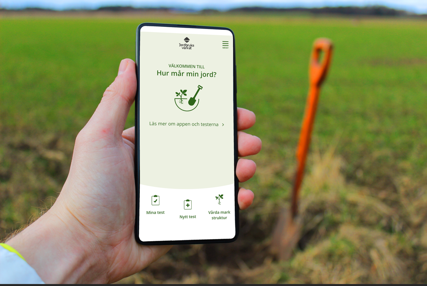 En hand som håller en mobil med appen Hur mår min jord. Fält och spade i bakgrunden.